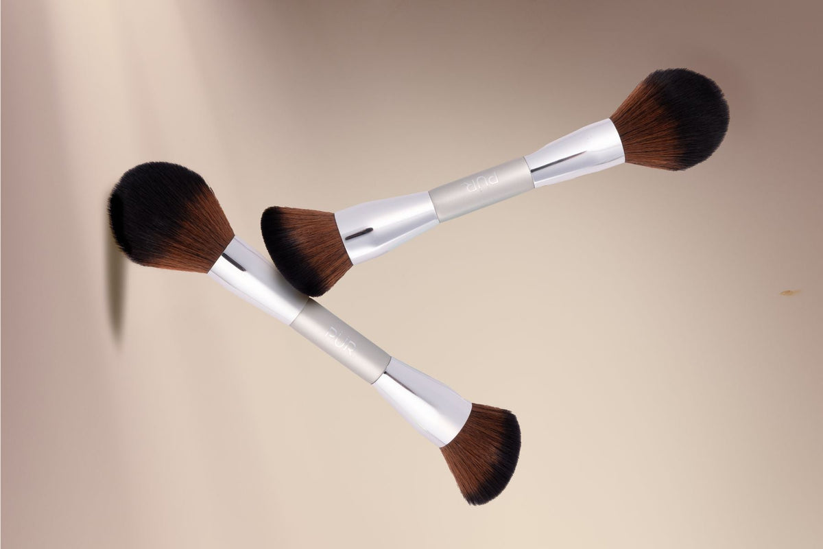 Two double-sided makeup brushes with silver handles and brown bristles arranged diagonally on a warm beige background.
