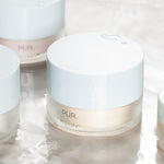 PÜR Tropical C Moisturizer jar with other skincare jars on a soft reflective surface, highlighting clean, minimal packaging and glossy white lids.
